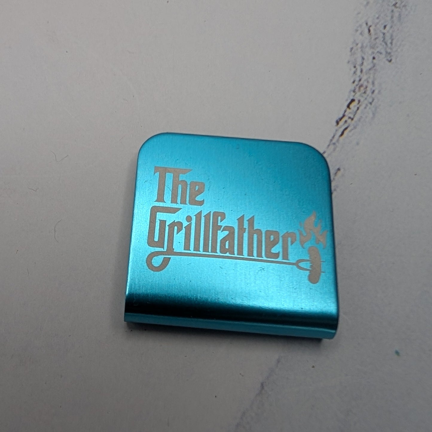 Teal metal hat clip with 'The Grillfather' text on a white background