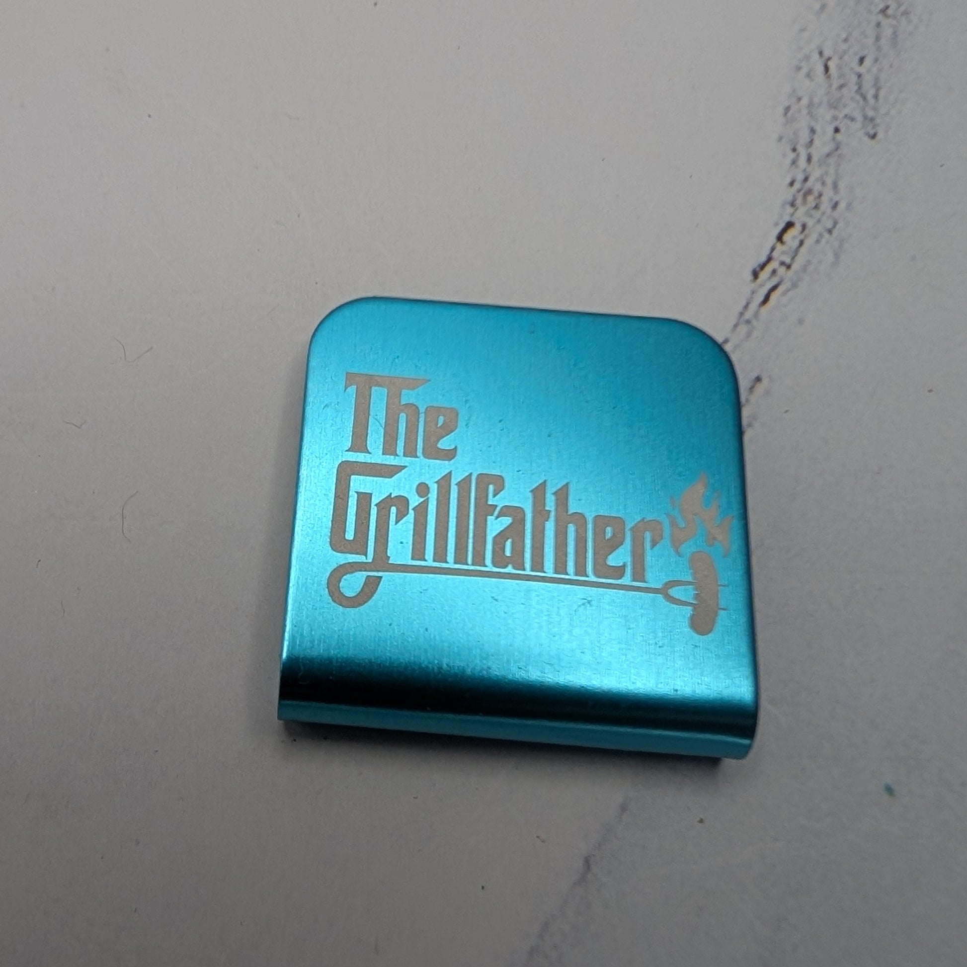 Teal metal hat clip with 'The Grillfather' text on a white background